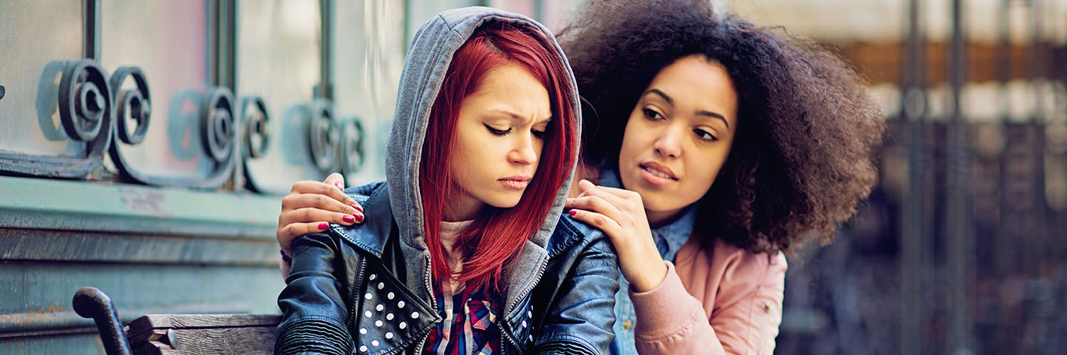 close up photo of two young women sitting on a bench, one woman is behind the other and has her hands on the shoulders of the other